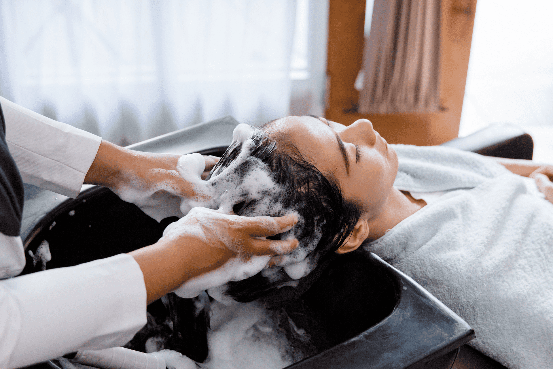 Person receiving a hair wash at a salon, lying back with eyes closed and towel around shoulders.